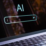 Laptop screen showing the text 'AI' with a glowing rounded search bar, conveying an AI/search concept on a dark display.