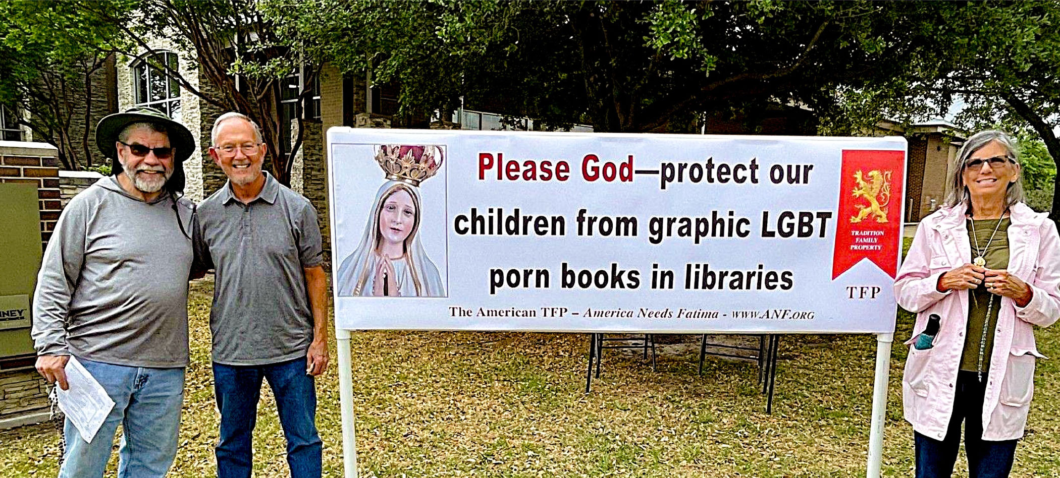 Three adults pose beside a large protest banner that reads, 'Please God—protect our children from graphic LGBT porn books in libraries,' outdoors.