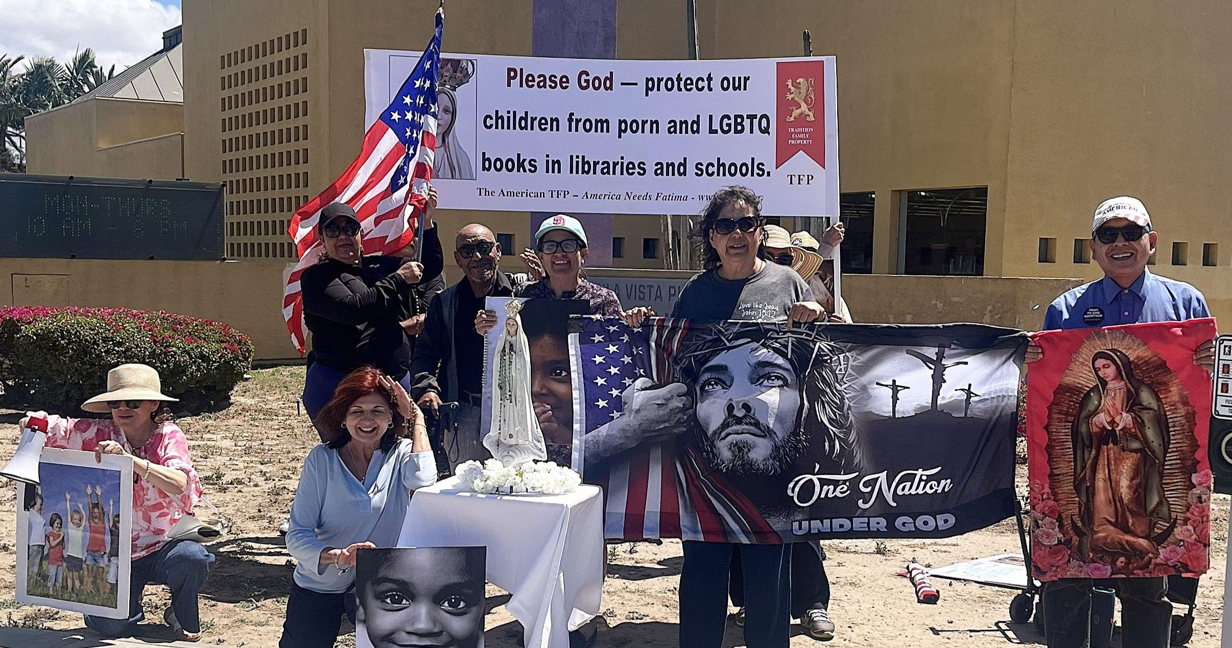 Group of people holding a banner urging to protect children from porn and LGBTQ content in libraries and schools, with an American flag nearby.