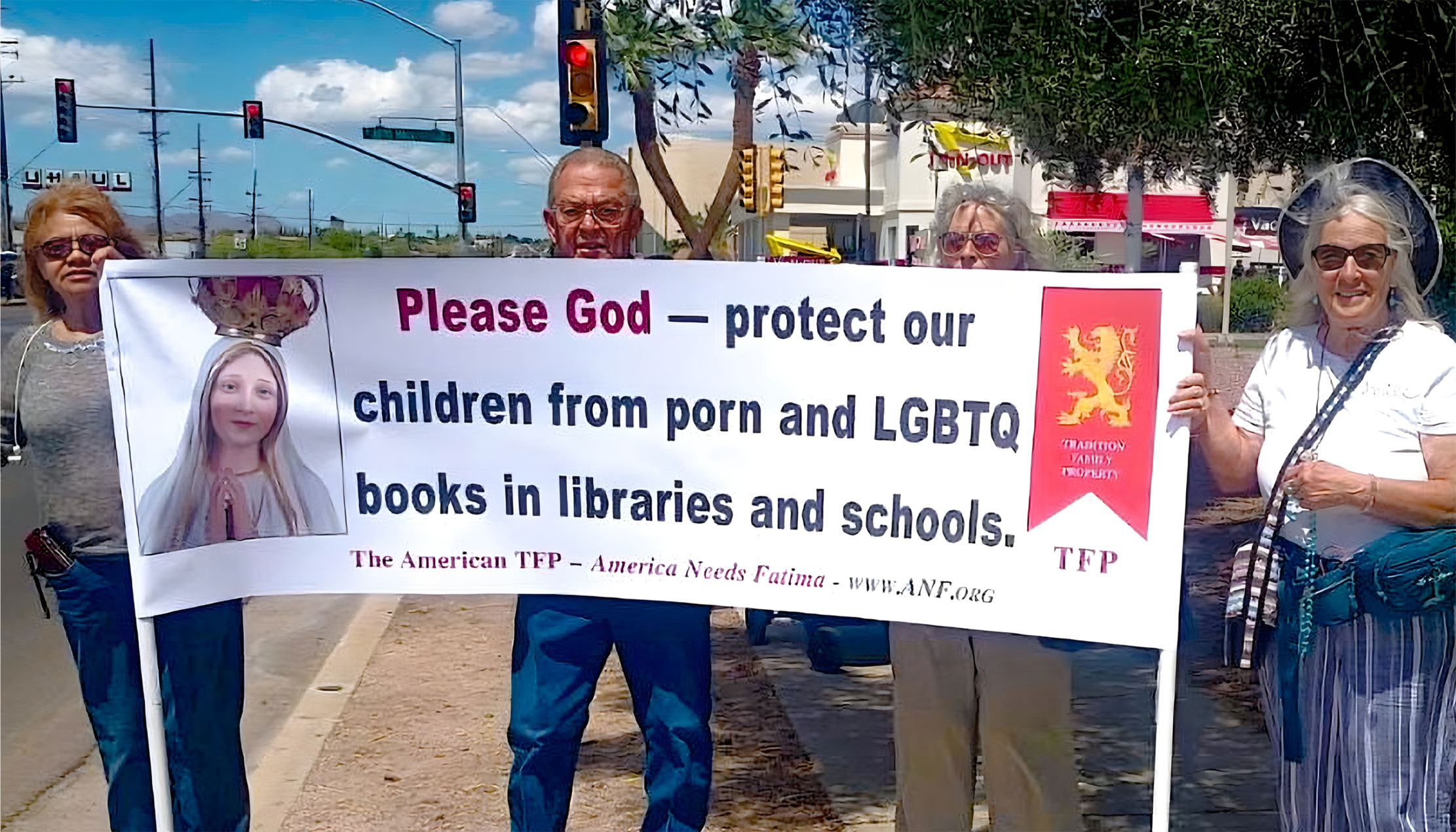 Group of protesters holding a large banner reading 'Please God—protect our children from porn and LGBTQ books in libraries and schools' on a street.