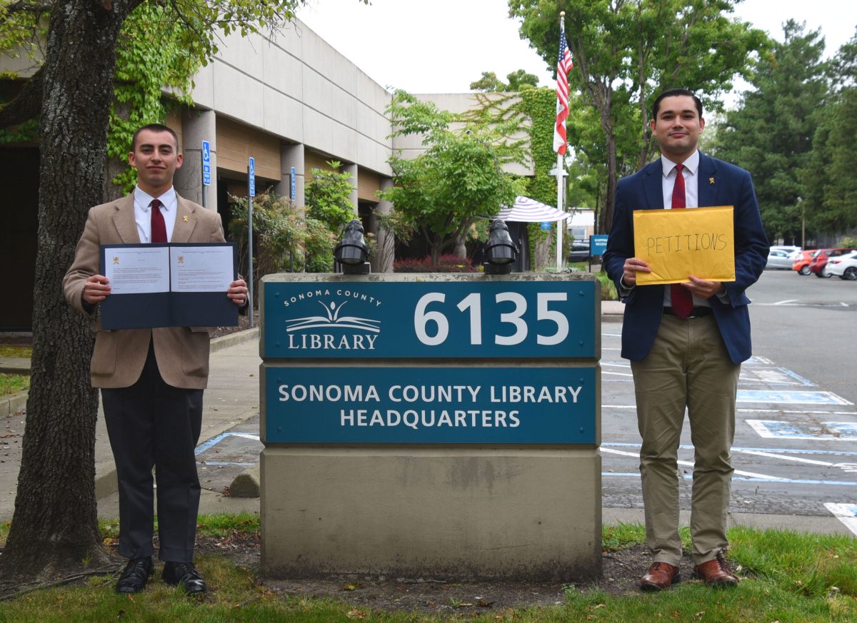 Two men in business attire stand on either side of a library sign outside the Sonoma County Library Headquarters, holding documents and a folder.