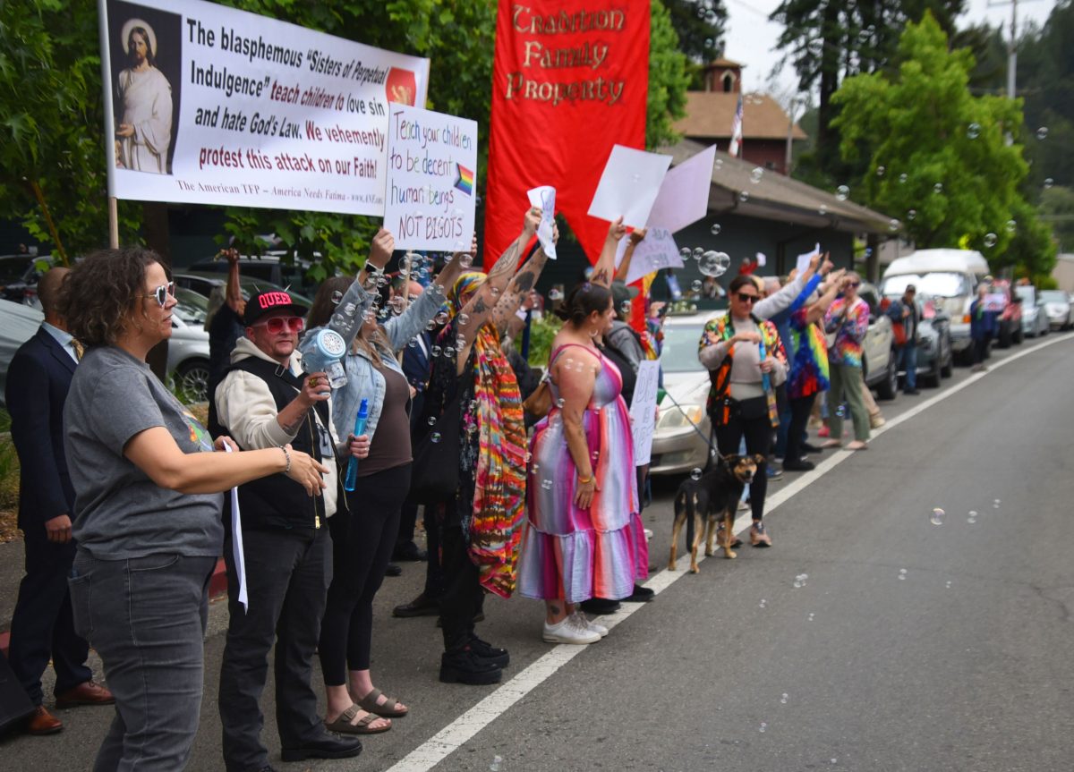 Line of protesters along a street holding signs and banners during a daytime demonstration, with bubbles floating in the air and colorful clothing.