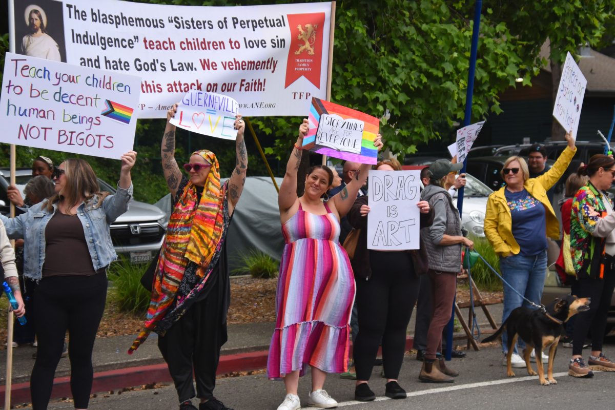 Group of protesters gathered on a street, holding handmade signs and a large banner in the background.