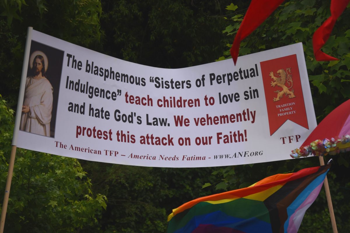 Large protest banner condemning a religious group, with a saint image on the left and a red emblem panel, and rainbow flags in the foreground.