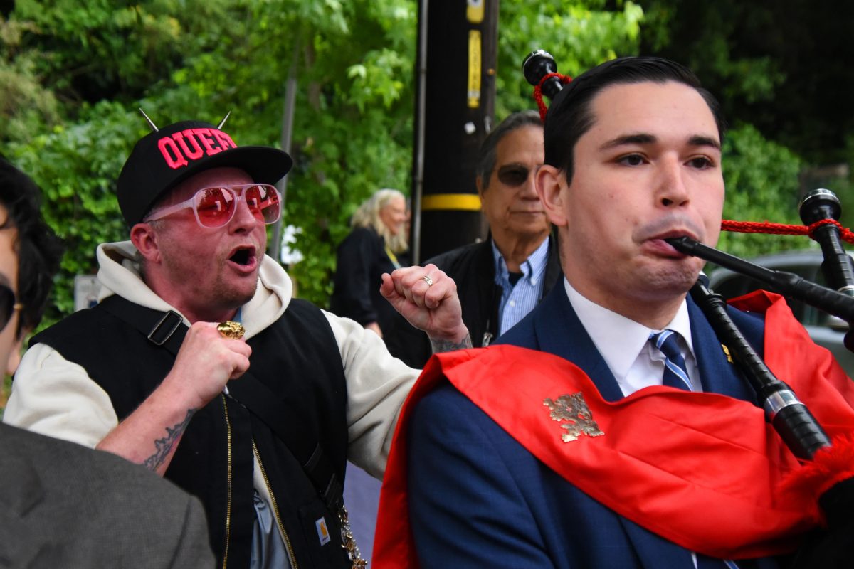 Man in a blue suit with a red cape playing bagpipes at a crowd event, the word 'QUEEN' visible on a cap worn by another man nearby.