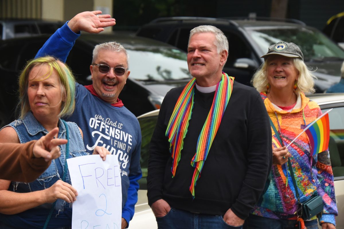Four adults at an outdoor event; a woman in a denim vest points toward the camera while two men stand beside her, one with a rainbow scarf and the other in a colorful jacket holding a rainbow flag.
