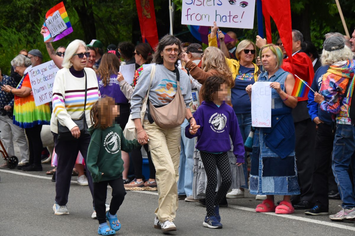 Crowd of diverse protesters marching, some holding handwritten signs and rainbow pride flags in a leafy outdoor setting.