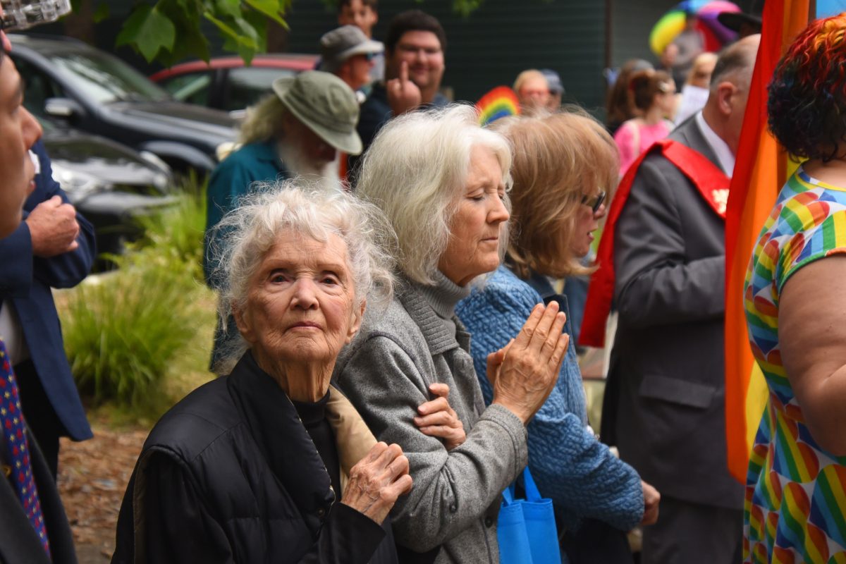 Group of people gathered outdoors for a celebration; in the foreground a gray-haired elderly woman prays with hands pressed together, others behind her, and bright rainbow-colored clothing visible on a participant to the right.
