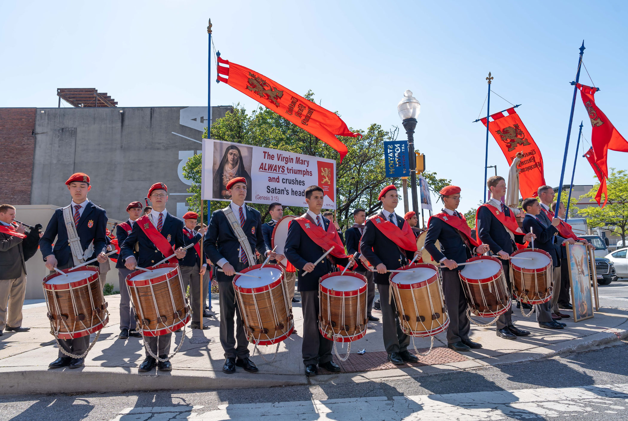 A line of young drummers in navy uniforms and red berets march with large drums, amid red banners and a banner about Mary and Genesis 3:15 in a street parade.