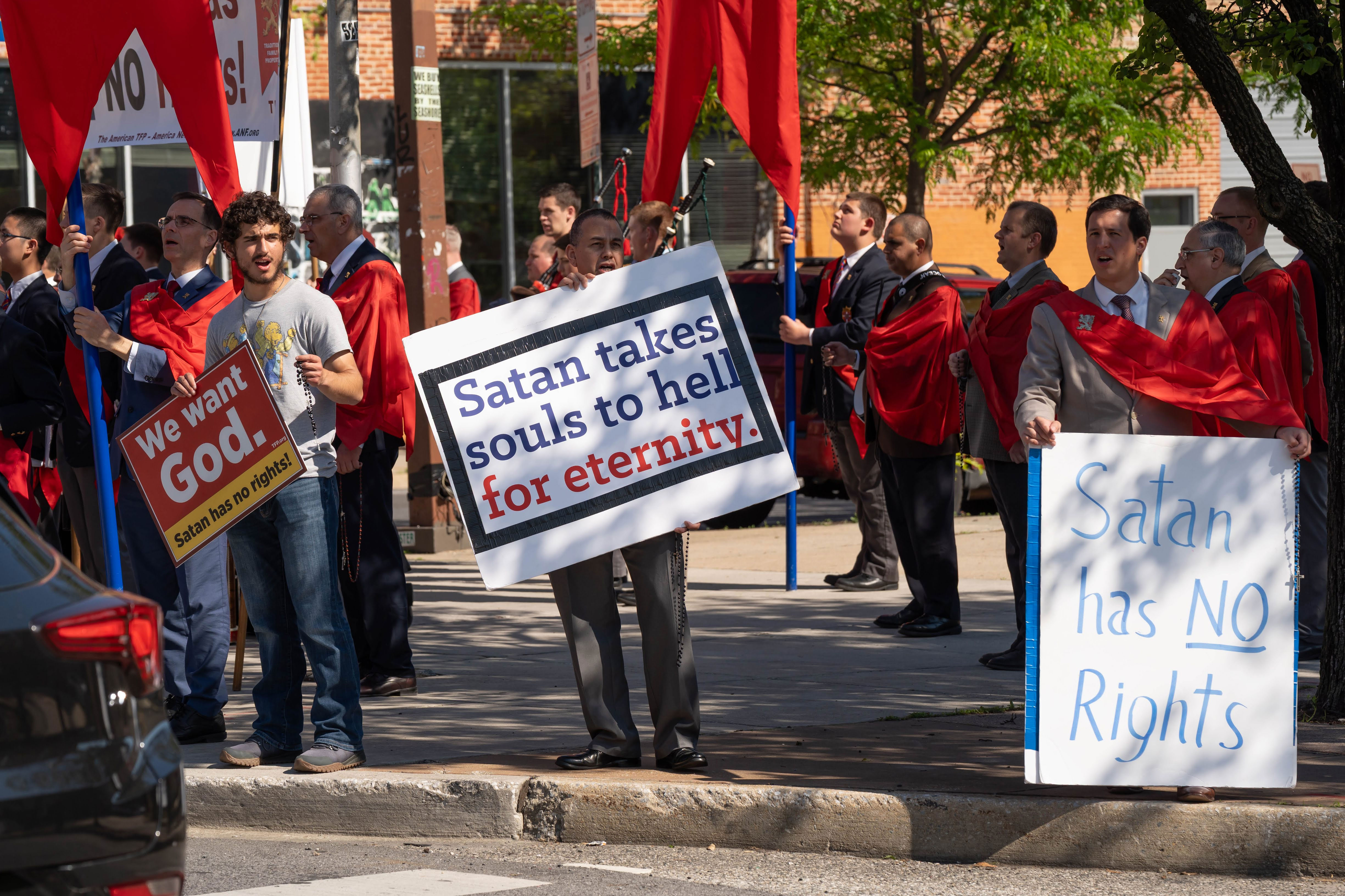 Protesters in red robes holding signs on a city street; central sign says 'Satan talks souls to hell for eternity.'