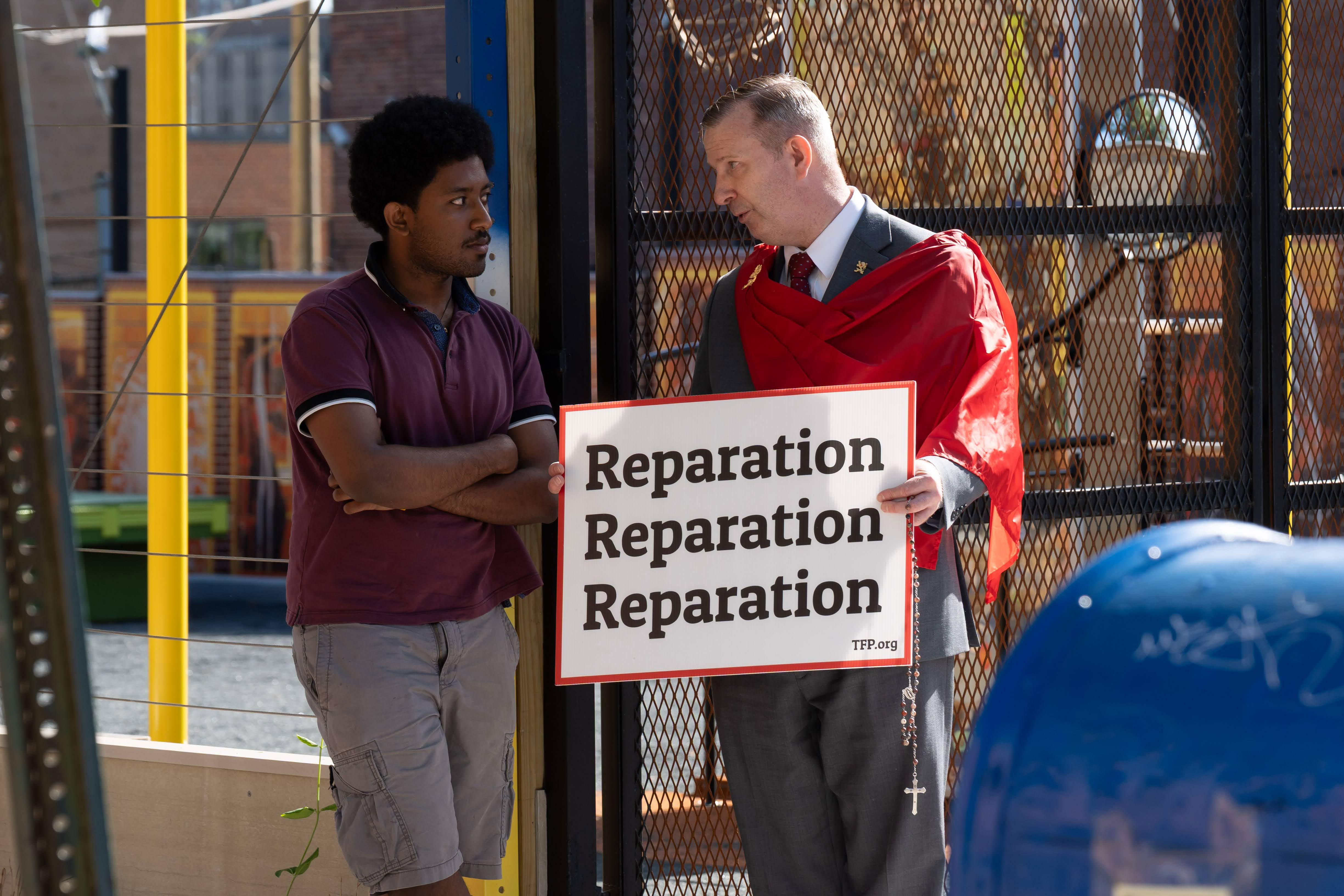 Two men stand beside a gated fence; one in a maroon polo with arms crossed, the other in a suit with a red cape, holding a 'Reparation' sign.