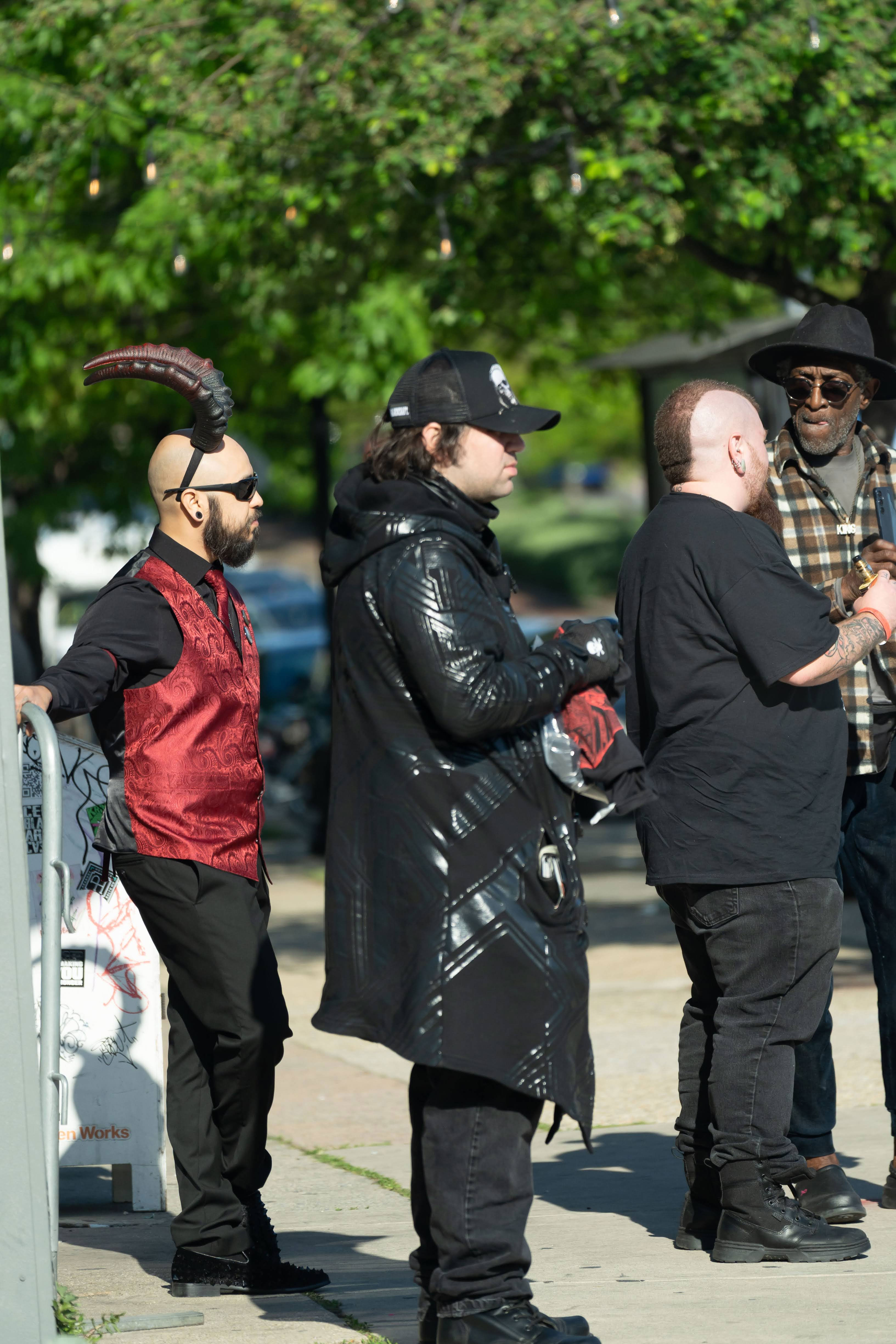 Group of men in costumes standing on a sidewalk, talking outdoors among trees.