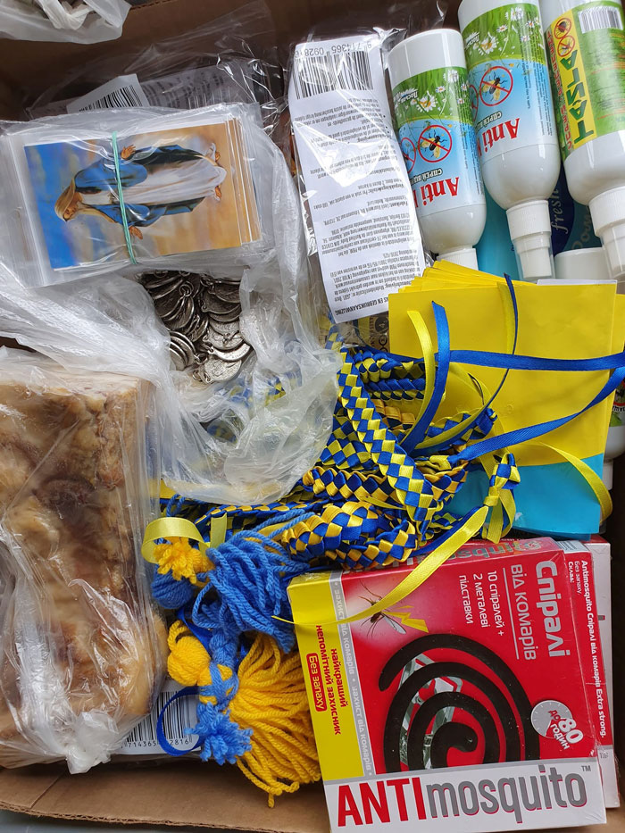Box of assorted items including yellow-blue woven cords, a red 'ANTImosquito' box, coins, a bar of soap, small religious cards, and white bottles with green labels.