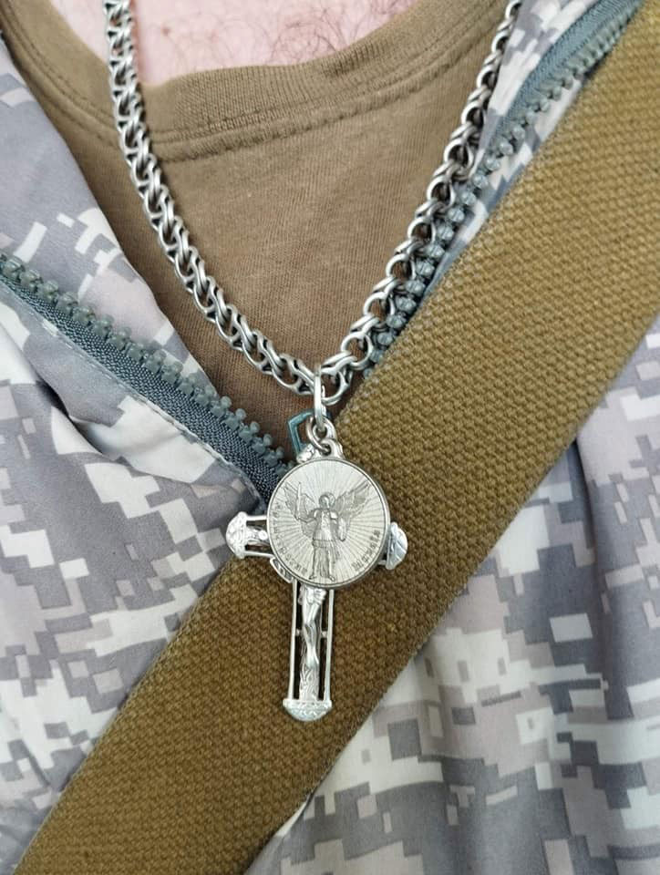 Silver chain necklace with a round medallion and a vertical crucifix pendant, worn over a brown shirt.