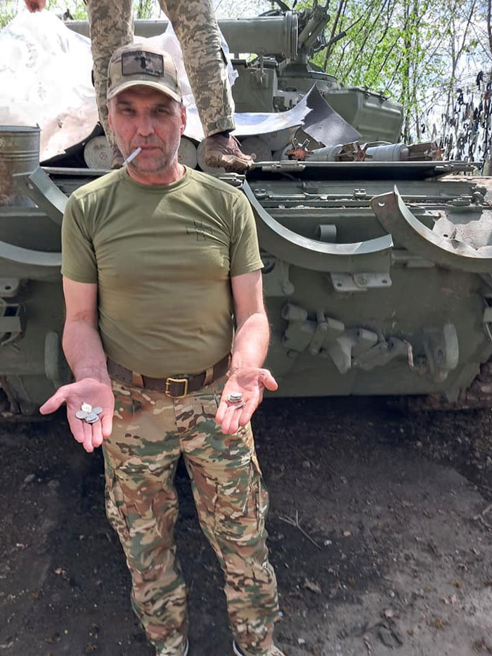 Man in olive-green shirt and camouflage pants stands in front of a military tank, holding several metal objects in his open palms with a cigarette in his mouth.
