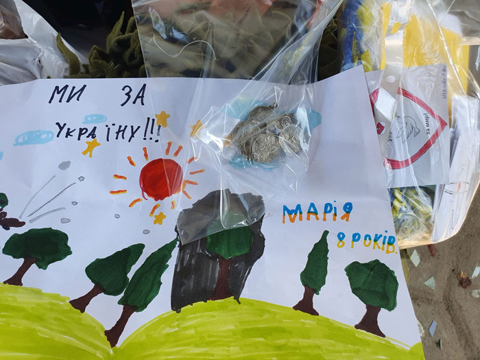 Colorful handmade Ukrainian poster with the slogan 'Ми за Україну!!!', a bright sun, green hills, trees, and a cave, signed 'Марія 8 років'. A small bag of coins rests on top.