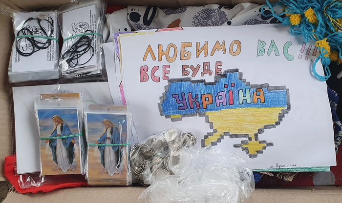 Display of a handmade Ukrainian patriotic poster with a blue‑yellow map and the words 'Любимо вас!!! Все буде Україна', next to bundles of religious cards in plastic sleeves and a pile of coins in a bag.