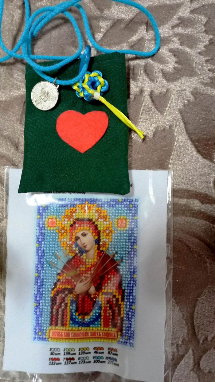Green fabric pouch with a red heart applique, blue cord necklace, and a small round silver charm resting on top. A beaded prayer-card icon lies below.