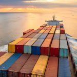 A cargo ship at sunset carrying stacked colorful shipping containers in the ocean.