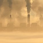 Factory complex with tall smokestacks releasing dark plumes into a hazy, yellow-tinted sky (air pollution)
