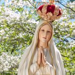 Statue of the Virgin Mary wearing a red crown, hands clasped in prayer, outdoors among blooming trees.