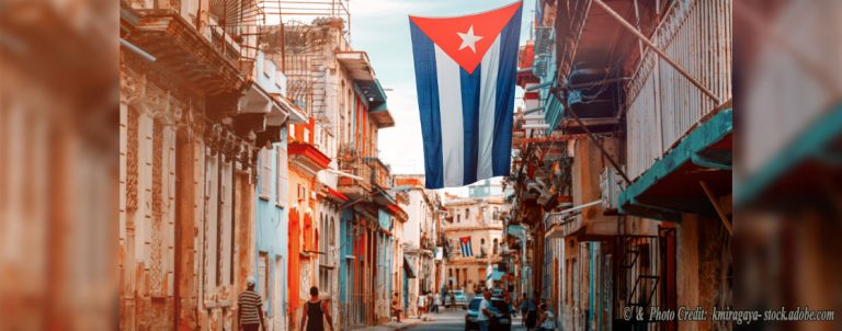 Cuban street lined with colorful colonial buildings and a large Cuban flag hanging above.