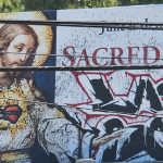 Billboard shows a Sacred Heart religious image on the left, with bold graffiti tagging across the right side.