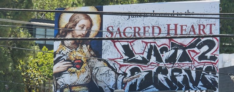 Billboard shows a Sacred Heart religious image on the left, with bold graffiti tagging across the right side.