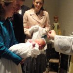 Priest in ornate robes blesses a newborn held by parents during a baptism ceremony.