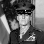 Black-and-white portrait of a female U.S. Army officer in dress uniform with ribbons and medals, standing before an American flag.