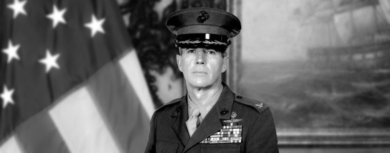 Black-and-white portrait of a female U.S. Army officer in dress uniform with ribbons and medals, standing before an American flag.