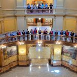 Dozens of people stand along the curved railing of a grand circular balcony inside a yellow-painted marble atrium, seen from above.