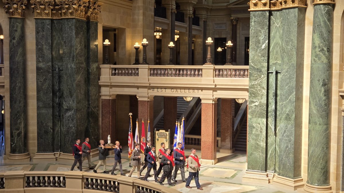 Ceremonial procession of uniformed men carrying flags through a grand marble hall with green pillars and ornate balconies.